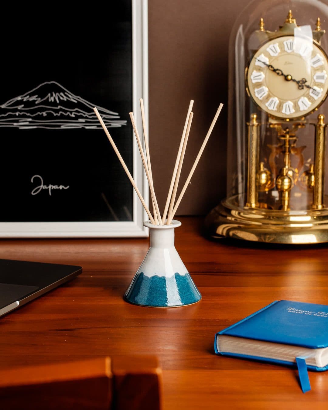 在胡桃木色的桌子上,中間擺著 FREESTONE 與陶拾光陶藝工作室合作的富士山造型陶瓷擴香瓶,擺在後面是黃銅色的發條桌鐘與白色畫框,畫框裡是極簡的富士山白色線條圖,桌上還有藍色的小冊子與銀色筆記型電腦的一角。